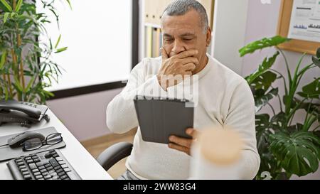 A mature hispanic man in a white sweater uses a tablet in a modern office, displaying concern or deep thought. Stock Photo