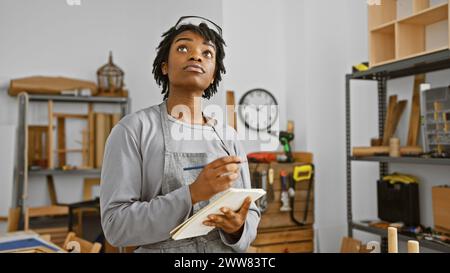 A thoughtful young african american woman with dreadlocks wears a grey apron in a woodworking studio, taking notes and planning a project. Stock Photo