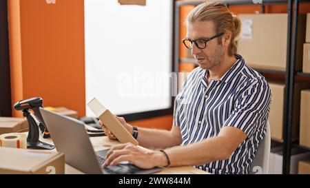 A man with long blonde hair is packaging products in a warehouse, using a laptop and barcode scanner amidst cardboard boxes. Stock Photo