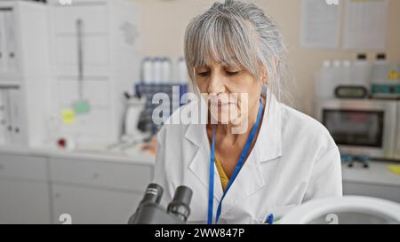 A middle-aged woman with grey hair working focused in a laboratory, examining a sample under a microscope. Stock Photo