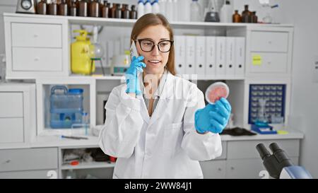 A caucasian woman scientist examines a petri dish in a laboratory whilst talking on the phone. Stock Photo