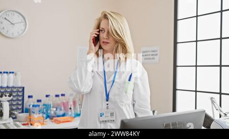 Blonde woman in lab coat on phone in laboratory, with badges, equipment, and daylight window. Stock Photo