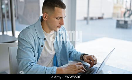 A focused young hispanic man using a laptop in a bright university library setting. Stock Photo