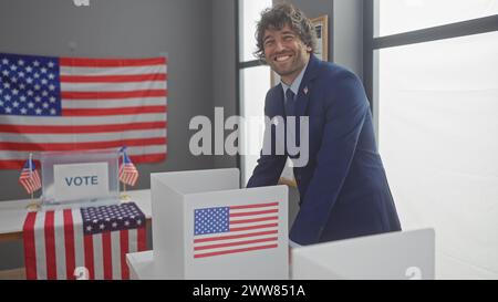 A smiling young hispanic man with a beard dressed in a suit stands by a voting booth with an american flag backdrop in an indoor setting. Stock Photo