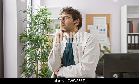 Pensive hispanic man with beard and glasses wearing wireless earphones in a modern office setting Stock Photo