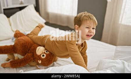 A playful blond boy enjoys time with a stuffed animal on a white bed in a bright bedroom. Stock Photo