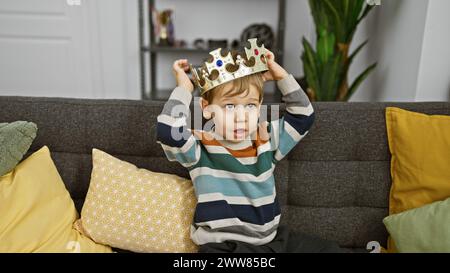 A surprised caucasian toddler boy with blond hair wearing a crown sits on a grey sofa indoors, surrounded by colorful cushions. Stock Photo