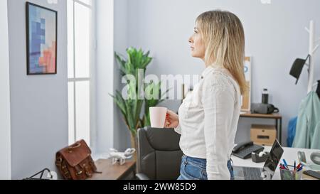 A middle-aged woman with blonde hair, standing thoughtfully in a modern office, holding a coffee cup. Stock Photo