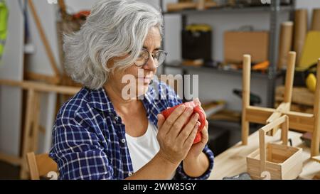 A mature woman inspects a wooden object with focus inside a well-equipped carpentry workshop. Stock Photo
