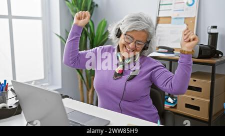 A joyful elderly woman with grey hair dances in an office, wearing headphones, glasses, and a purple sweater. Stock Photo