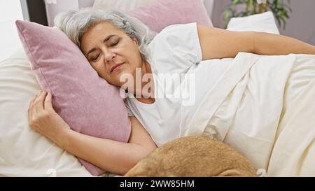 Grey-haired woman sleeping in bed with a pet dog in a cozy home bedroom. Stock Photo