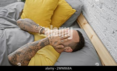 Tattooed young man laying on bed in a modern bedroom interior, covering his face with hands showing emotion. Stock Photo