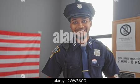 Smiling african american policeman with 'i voted' sticker in uniform posing in front of us flag indoors. Stock Photo