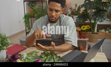 A focused black man calculating costs with a phone in a flower shop surrounded by green plants and a laptop. Stock Photo