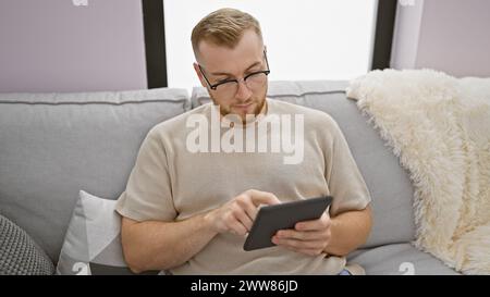 A young bearded man in glasses intently uses a tablet on a cozy sofa in a modern living room. Stock Photo