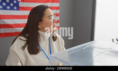 A thoughtful hispanic woman stands near an american flag in an indoor setting, possibly at an electoral event. Stock Photo
