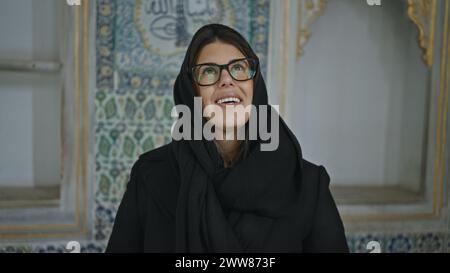 A joyful woman in glasses wearing a hijab stands in an ornate turkish palace, featuring intricate tile work. Stock Photo