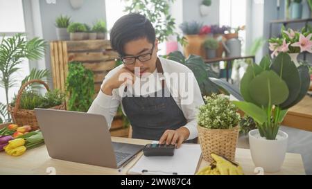 A focused asian man calculating finances on a calculator in a flower shop filled with various plants. Stock Photo
