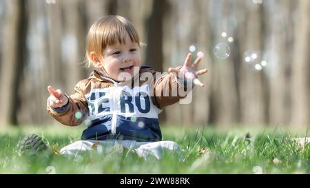 Happy cute smiling baby boy sitting on green grass outdoor playing with soap bubbles. Concept of love, care, parenthood Stock Photo