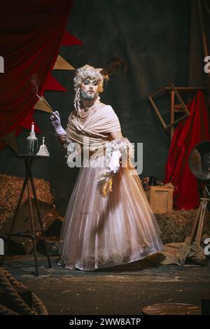 Drag queen in white dress and bright makeup over dark retro circus backstage background. Unusual show Stock Photo