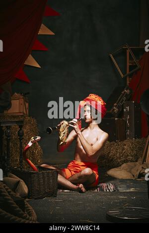 Young man, charmer in turban playing flute with cobra emerging from basket over dark retro circus backstage background. Stock Photo