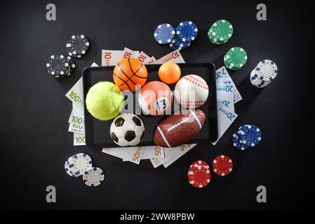 Online sports betting concept with various sports balls, money and chips on mobile devices isolated in the center on dark background. Top view. Stock Photo