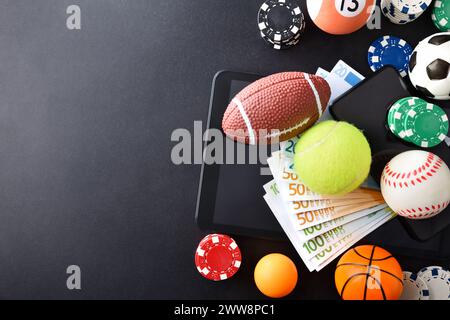 Online sports betting concept with various sports balls, money and chips on mobile devices isolated on the right on a black table. Top view. Stock Photo