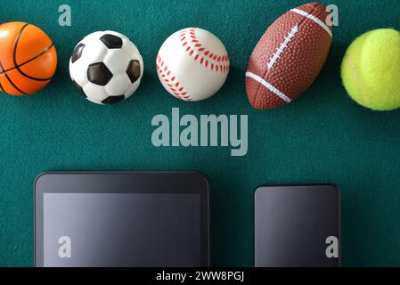 Concept of sporting events and technology with tablet and smartphone lined up on green felt with balls of various sports in a row. Top view. Stock Photo