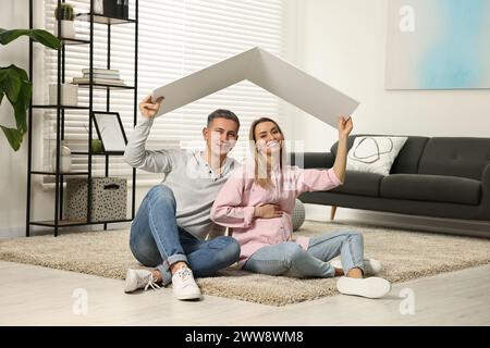 Young family housing concept. Pregnant woman with her husband sitting under cardboard roof on floor at home Stock Photo