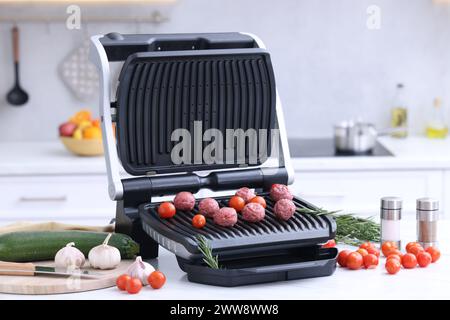 Electric grill with meatballs, tomatoes and vegetables on white table in kitchen Stock Photo