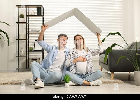 Young family housing concept. Pregnant woman with her husband sitting under cardboard roof on floor at home Stock Photo