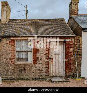 Atmospheric old derelict red brick shed with peeling paint door and windows on a grey sky day. Dorset Coast. Historical. Stock Photo