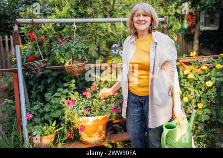 An elderly smiling woman waters flowers in hanging pots from a watering can. An aged positive woman in casual clothes takes care of plants. A beautifu Stock Photo