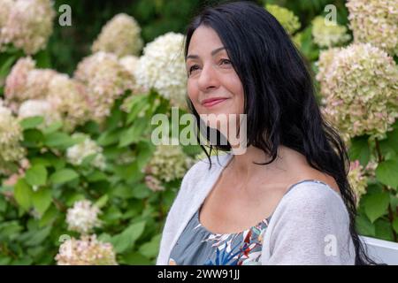 Serenely poised against a garden, flower, backdrop, a woman exudes confidence and contentment, embodying the grace of embracing midlifes struggles wit Stock Photo