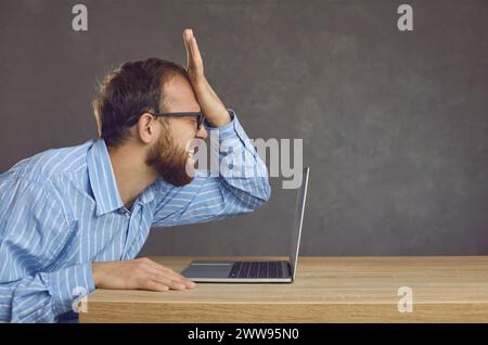 Man makes a stupid mistake while using his computer and slaps himself on his face Stock Photo