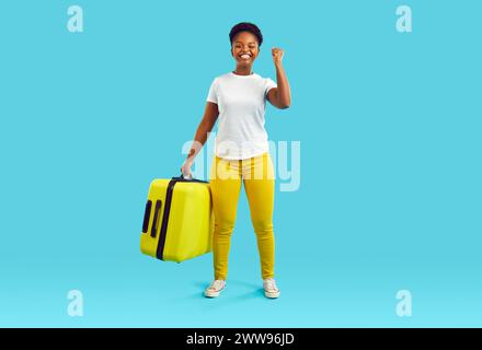 Happy woman carry on huge yellow suitcase on blue background. Lady with smile and victory gesture. Stock Photo
