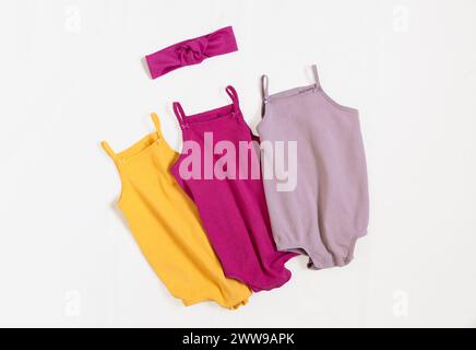 Cute kids summer outfit. Three baby vest bodysuit yellow, pink, lilac color with head accessories. Top view, flat lay on white. Stock Photo