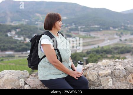 Adult active woman travel with backpack relaxes sit with water bottle on hill after hiking up, look at camera. Healthy lifestyle in retirement. Adventure seeks woman treks enjoys freedom and vacation. Stock Photo
