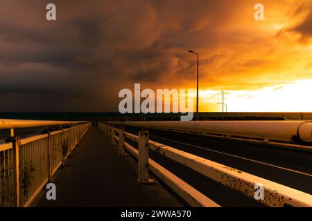 The dark silhouette of a bridge against a fiery sunset over a quiet harbor. Stock Photo