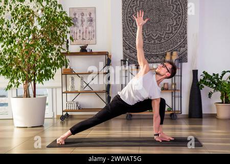 Full body of fit barefoot guy in activewear and eyeglasses practicing revolved lunge yoga pose on mat during practice session in modern studio with po Stock Photo