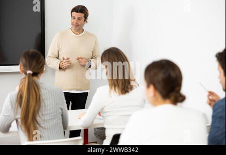 Male business coach explaining topic while standing against group of people sitting at desks in front of him Stock Photo