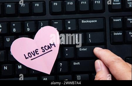Pink heart with love scam text placed on a computer keyboard while a finger presses the enter key Stock Photo