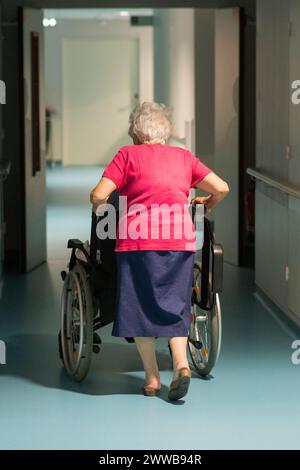 EHPAD - Elderly woman pushing her resident husband in a wheelchair down a hallway. Stock Photo