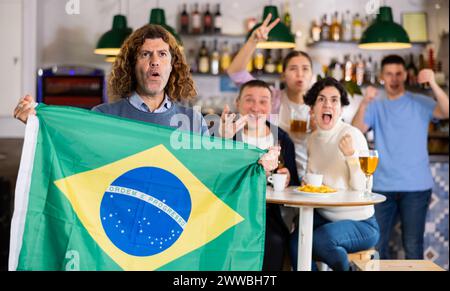 Company of emotional young adult fans supporting sports team of Brazil with state flag while resting with beer in bar Stock Photo