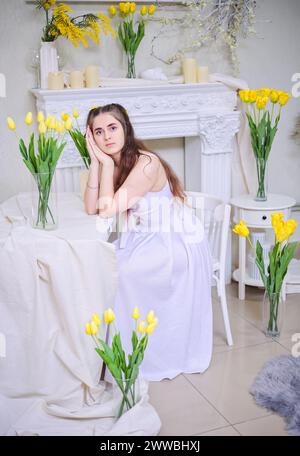 A beautiful young girl in a white dress and with long wavy hair against a background of yellow tulips. Stock Photo