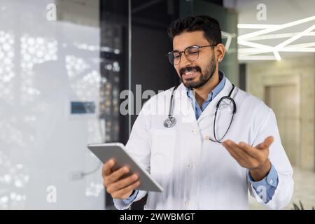 A doctor is holding a tablet and smiling. He is wearing a white lab coat and glasses. The tablet is open to a page with a picture of a person Stock Photo