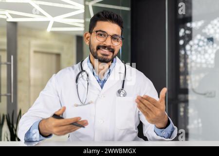 A doctor is smiling and holding a clipboard. He is wearing a white lab coat and glasses. The clipboard has a pen on it Stock Photo