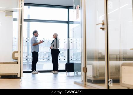 Mature businessman talking to colleague standing near window in office Stock Photo