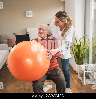 Home caregiver assisting senior man doing exercise with fitness ball in living room Stock Photo