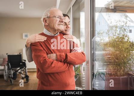 Smiling home caregiver embracing senior man looking through window Stock Photo
